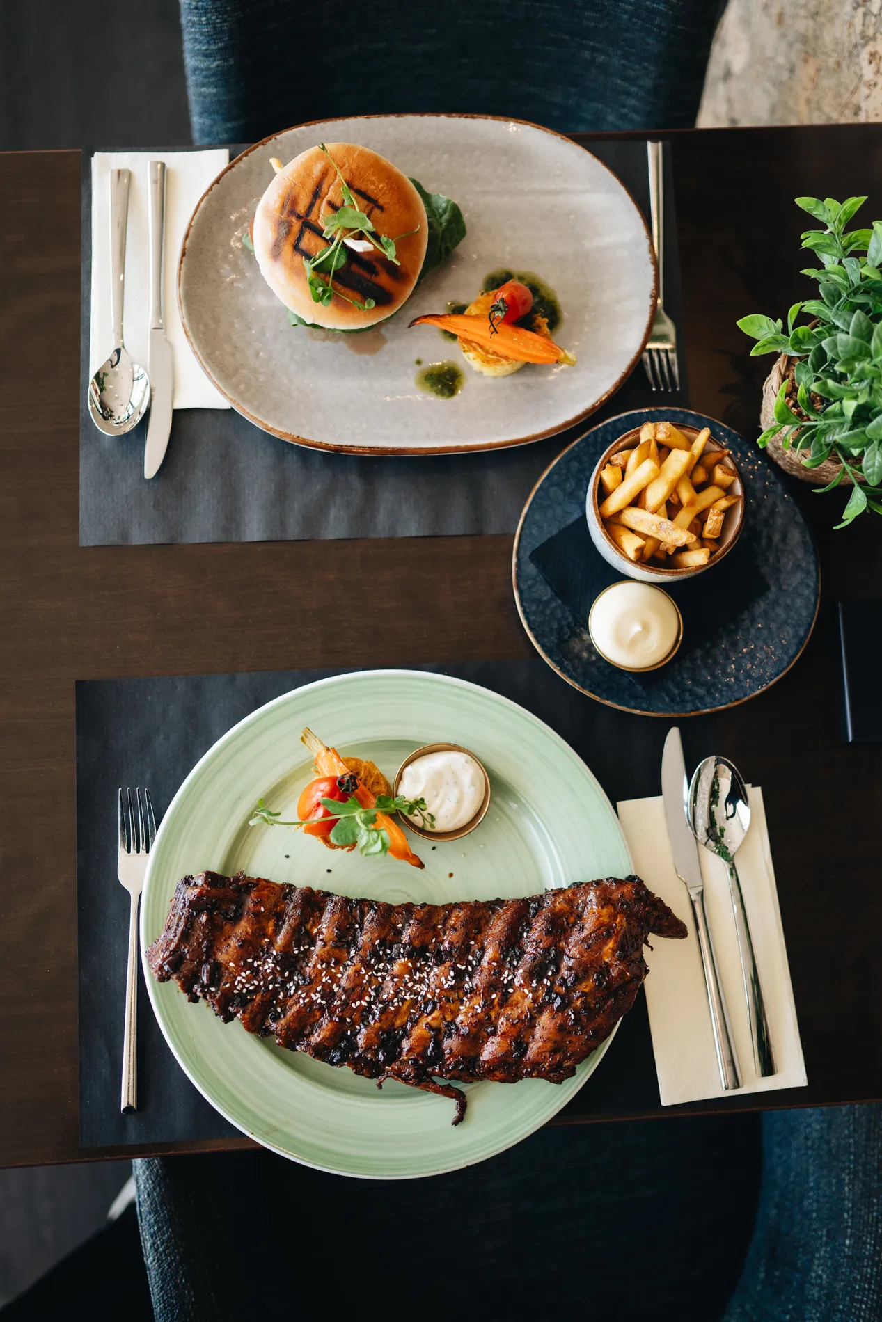 Verticaal bovenaanzicht van een houten tafel met onderaan een grote streng spareribs op een lichtgroen bord en bovenaan een hamburger met gegrilde strepen op het broodje, geflankeerd door een schaaltje frites.